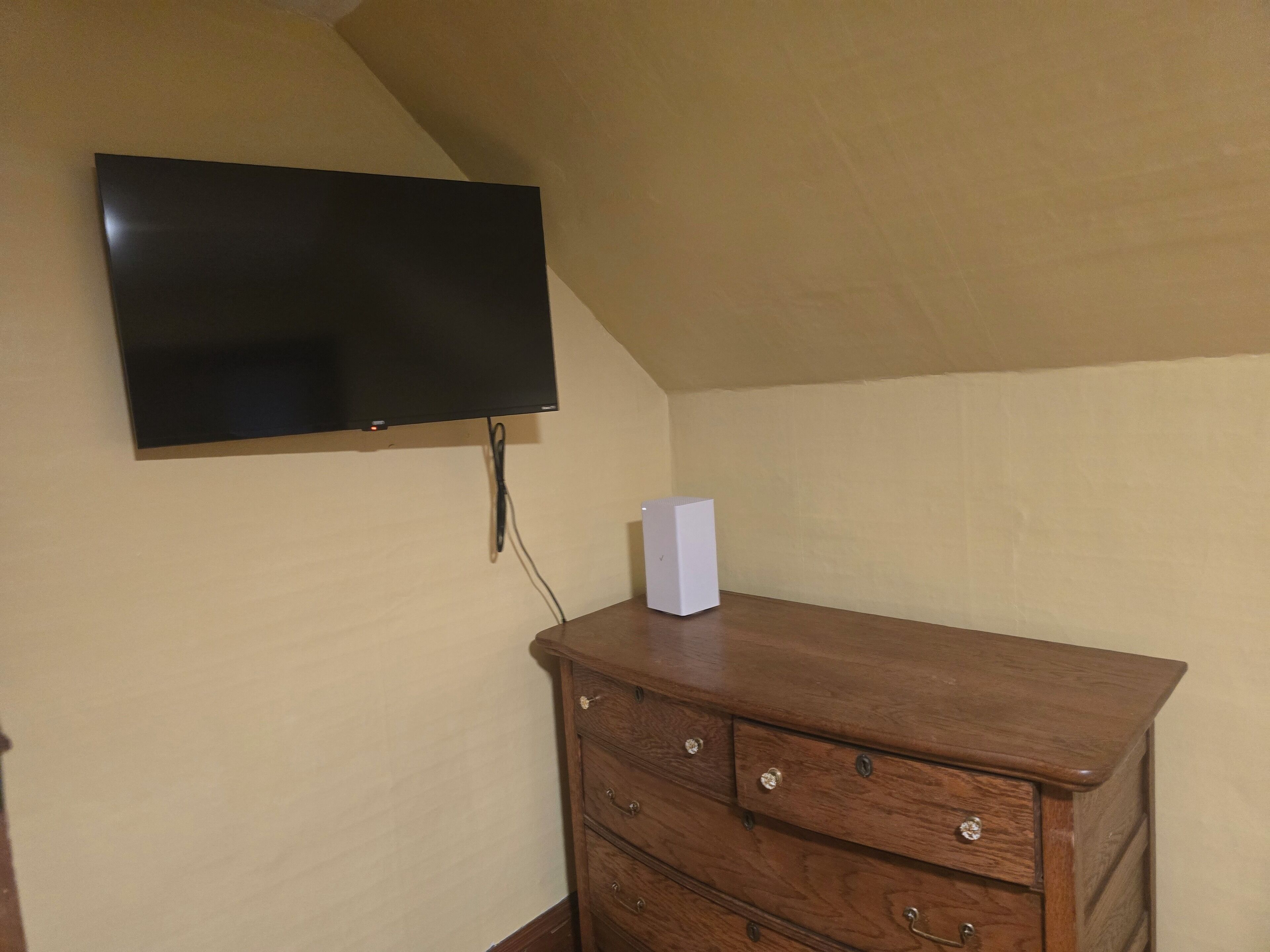 A room with a flat-screen TV mounted on the wall, a wooden dresser with drawers, and a white box on top.