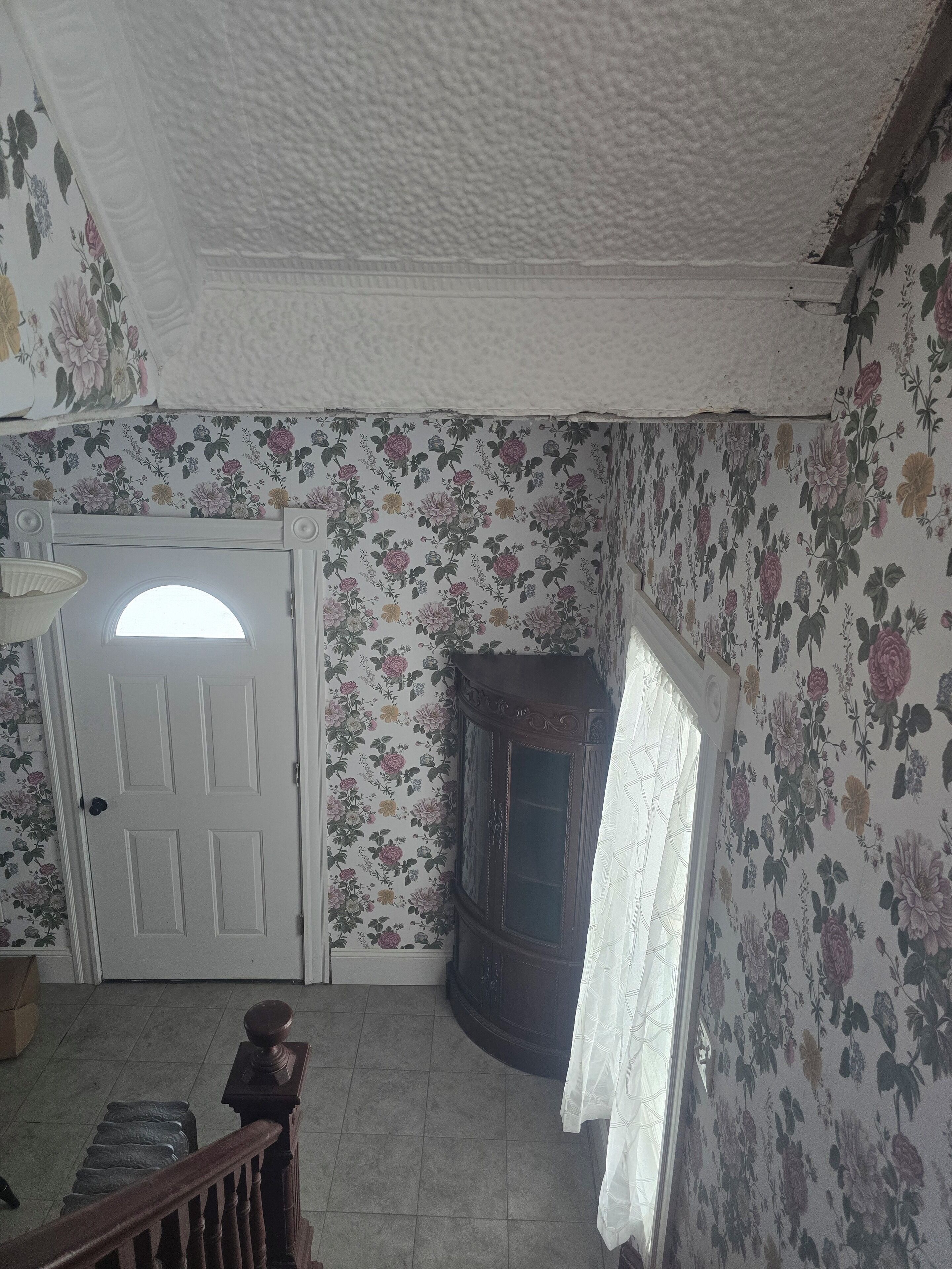 A room with floral wallpaper, a white door, a dark wooden cabinet, and a staircase with a wooden railing.