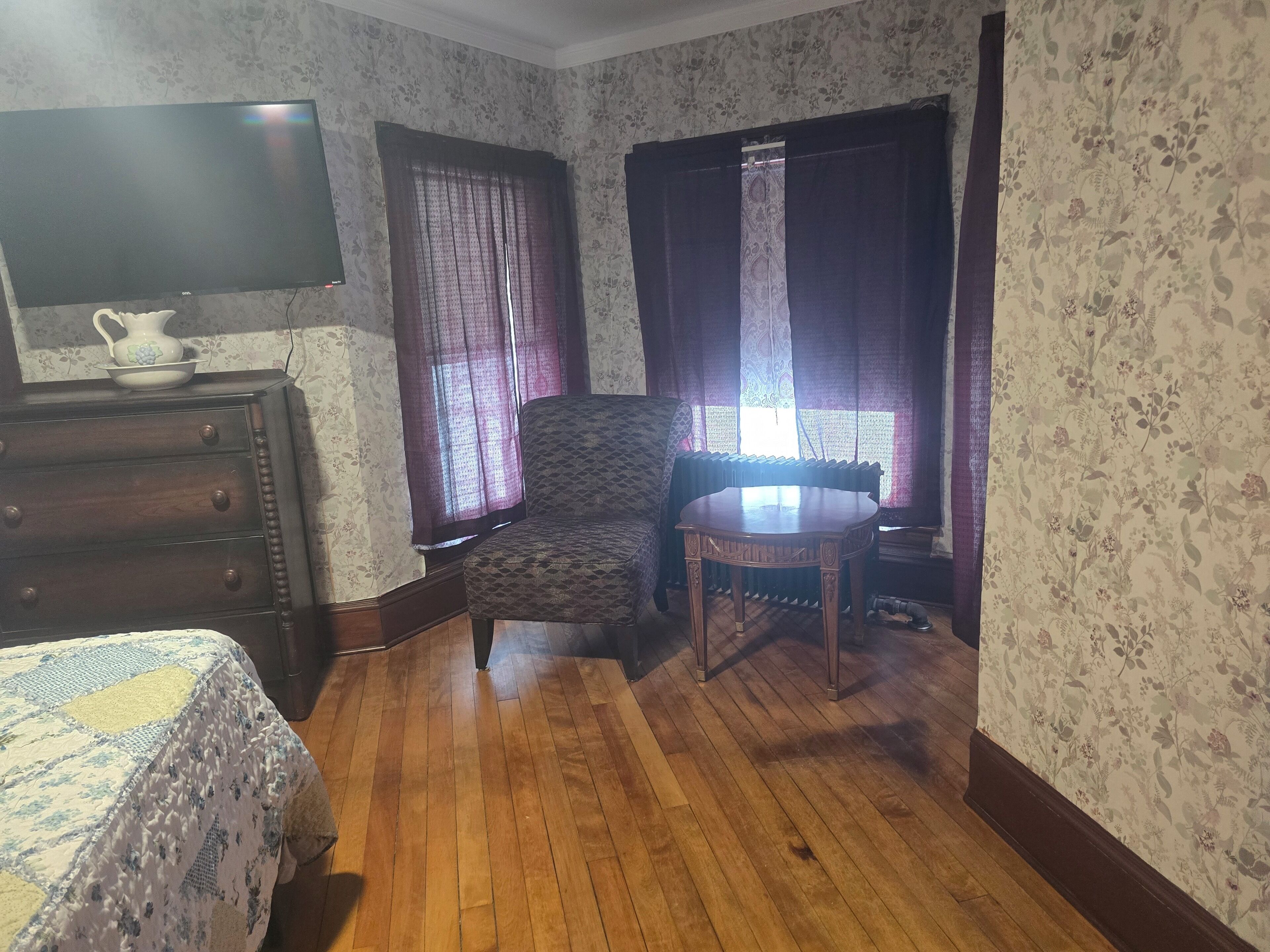 Una habitación con piso de madera, una cama con colcha estampada, un aparador, una silla, una mesa y una ventana con cortinas.