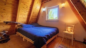 Un dormitorio acogedor en el ático, con techo inclinado de madera, una cama individual con manta azul, una mesita con lámpara y una ventana con vistas a árboles.