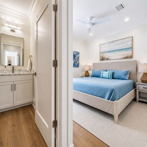 A bedroom with a bed, bedside tables, a ceiling fan, a painting, and a bathroom with a mirror and cabinets. - Newer Luxury 4-bedroom 4-bath cottage with community pool 2 minute walk to beach (Gulf Shores)