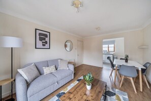 Ein Wohnzimmer mit grauem Sofa, einem Couchtisch mit Pflanze, einem Essbereich mit weißem Tisch und grauen Stühlen sowie einer Küche im Hintergrund.