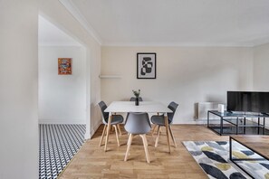 Ein modernes Wohnzimmer mit Essbereich, ausgestattet mit einem weißen Tisch, grauen Stühlen, einem Glastisch und einem Flachbildfernseher.