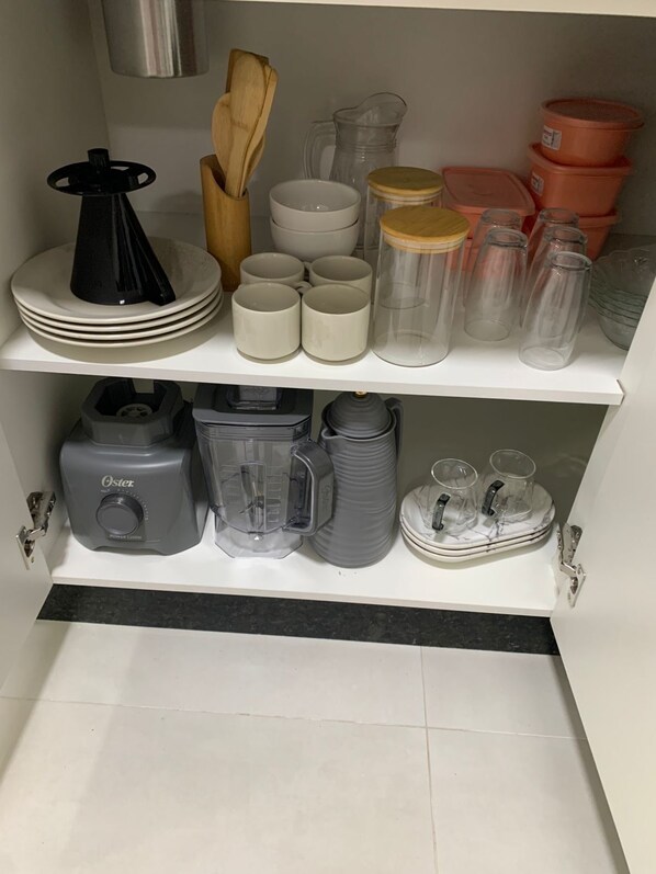 Een keukenkastje met diverse keukengerei, waaronder borden, mokken en glazen containers.