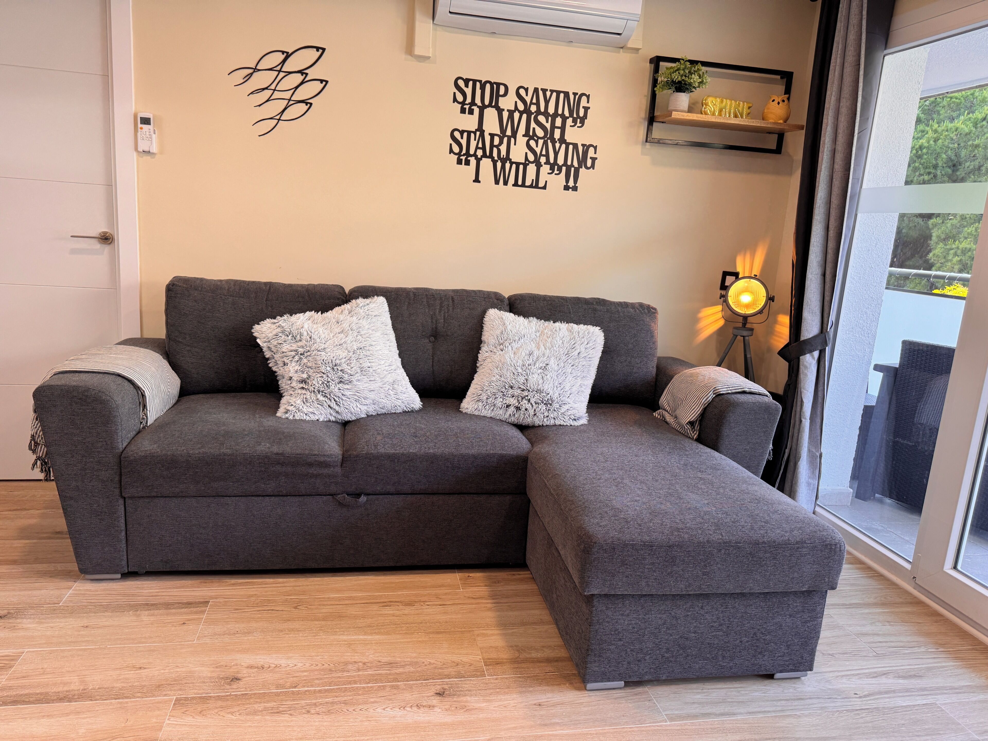 Een grijze modulaire bank met zachte witte kussens, een wandairconditioner, een inspirerende quote, een klein plankje met decoratieve objecten en een raam met uitzicht op groen.