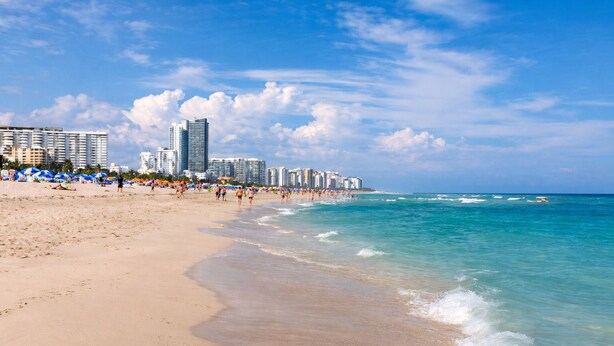 Uma praia com paisagem urbana ao fundo, incluindo edifícios altos e céu claro.