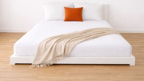 Uma cama com roupa de cama branca, um cobertor bege e um travesseiro laranja.