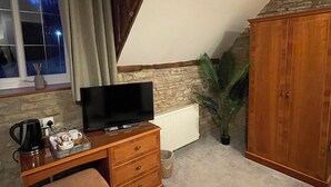 A room with a wooden desk, a television, a window with curtains, a radiator, a potted plant, and a wardrobe. - The Bateman Arms (Shobdon)