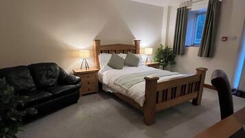A bedroom with a bed, a wooden headboard, a nightstand, a lamp, a plant, a window with curtains, and a couch.