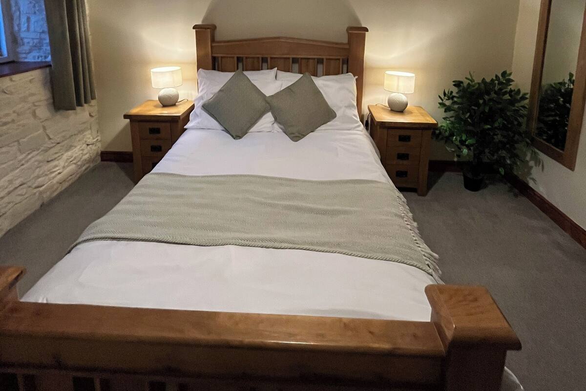 A bedroom with a wooden bed, two bedside tables with lamps, a ceiling light, a stone wall, a window with curtains, and a potted plant.