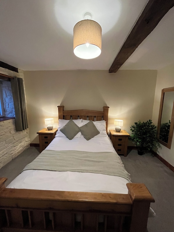 A bedroom with a wooden bed, two bedside tables with lamps, a ceiling light, a stone wall, a window with curtains, and a potted plant. - The Bateman Arms (Shobdon)
