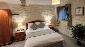 A bedroom with a stone wall, a wooden headboard, a bed with white linens, two bedside lamps, a window with curtains, and a framed picture on the wall. - The Bateman Arms (Shobdon)