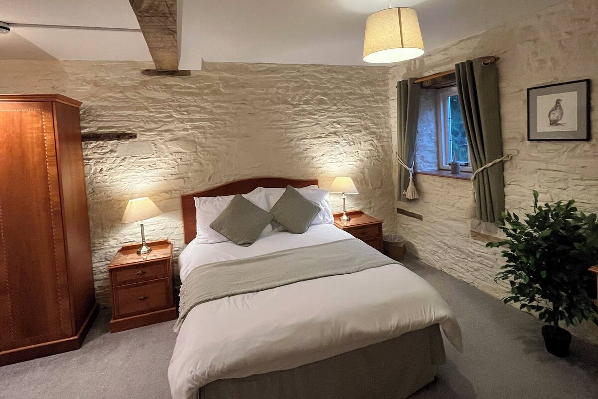 A bedroom with a stone wall, a wooden headboard, a bed with white linens, two bedside lamps, a window with curtains, and a framed picture on the wall.