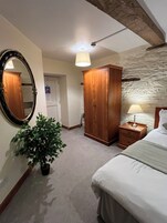 Un dormitorio con una cama, un armario de madera, una mesita de noche con lámpara, una planta en maceta, un espejo y una puerta.
