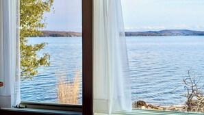 Ein Fenster mit transparenten weißen Vorhängen, Blick auf einen ruhigen See und eine entfernte Uferlinie mit Bäumen und Hügeln.