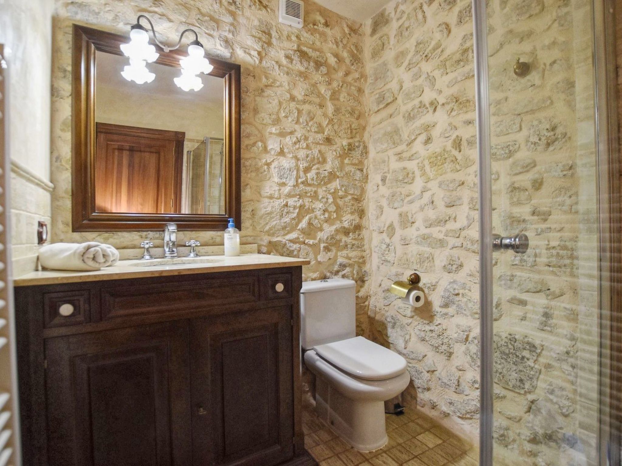 A bathroom with a stone wall, a wooden vanity with a mirror, a toilet, and a shower area.