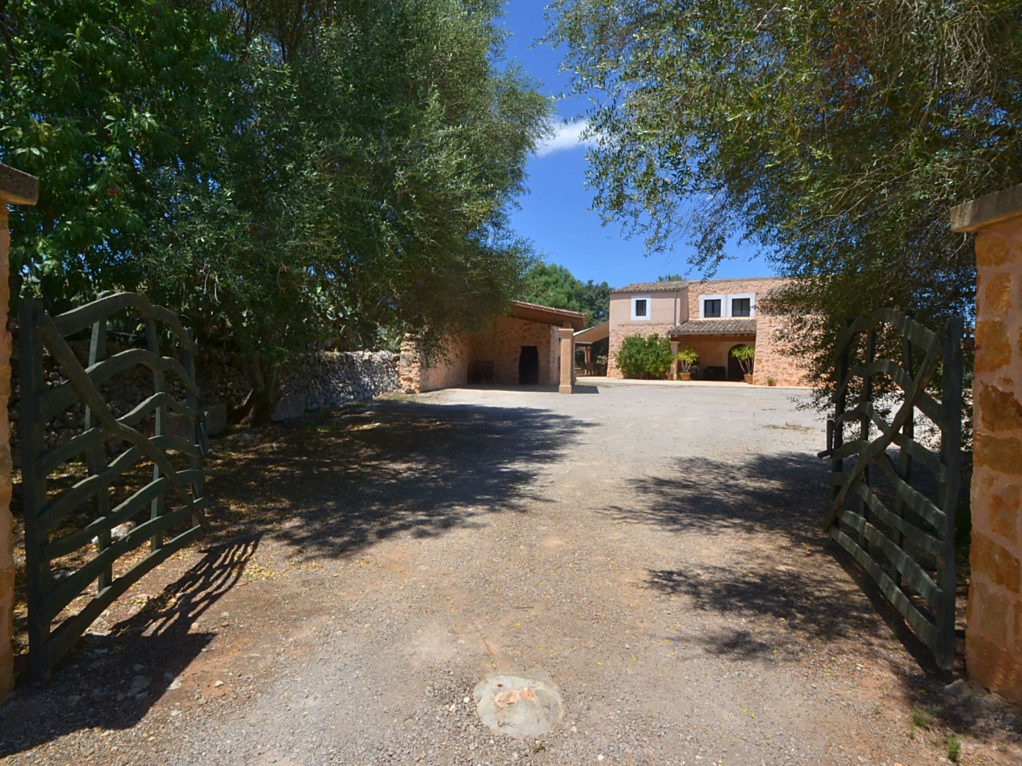 A gravel driveway leading to a stone building with a tiled roof, surrounded by trees.
