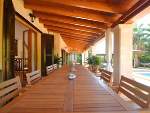 A covered outdoor dining area with wooden tables and benches, potted plants, and a view of a pool and greenery. - Wunderschön restauriertes Landhaus by Interhome (Son Sardina)