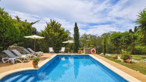 Una piscina rectangular con borde enlosado, rodeada de sillones y sombrillas, ubicada en un jardín ajardinado con diversas plantas y árboles.