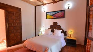 Ein Schlafzimmer mit Holztür, einem Bett mit weißer Bettwäsche, zwei Nachttischen mit Lampen und einem farbenfrohen Gemälde an der Wand.