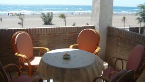 Een balkon met rieten stoelen en een tafel, met uitzicht op een strand met mensen en palmbomen.