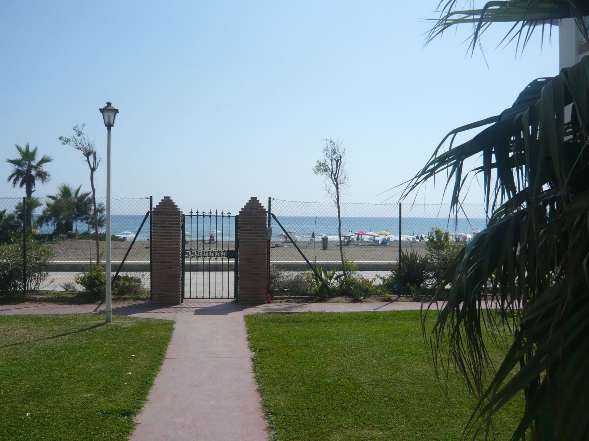 Un sendero que lleva a una playa con una reja metálica, palmeras y un farol.