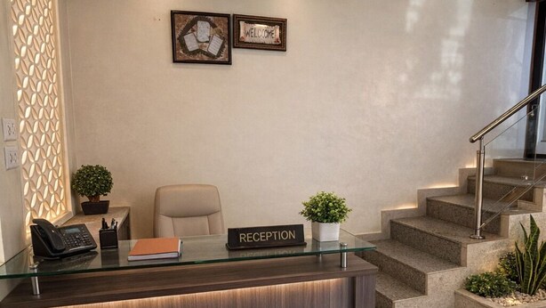 A reception area with a dark wood reception desk, a glass top, and a 'RECEPTION' sign. There are two framed welcome signs on the wall, a staircase to the right, and a decorative wall panel on the left. - Hotel rosewood plaza - Mahabaleshwar (Mahabaleshwar)