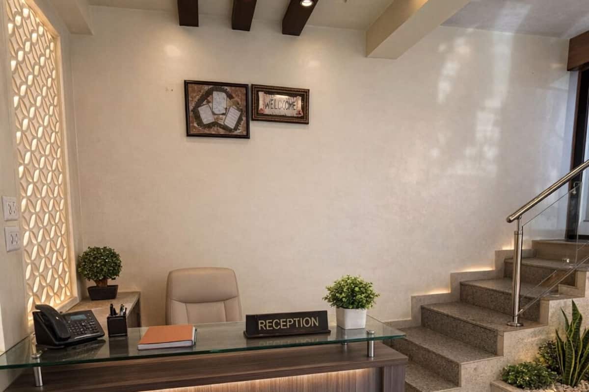 Ein Empfangsbereich mit dunklem Holzbüro, einer Glastischplatte und einem Schild mit der Aufschrift 'RECEPTION'. An der Wand befinden sich zwei gerahmte Willkommensschilder, eine Treppe zur Rechten und eine dekorative Wandverkleidung zur Linken.