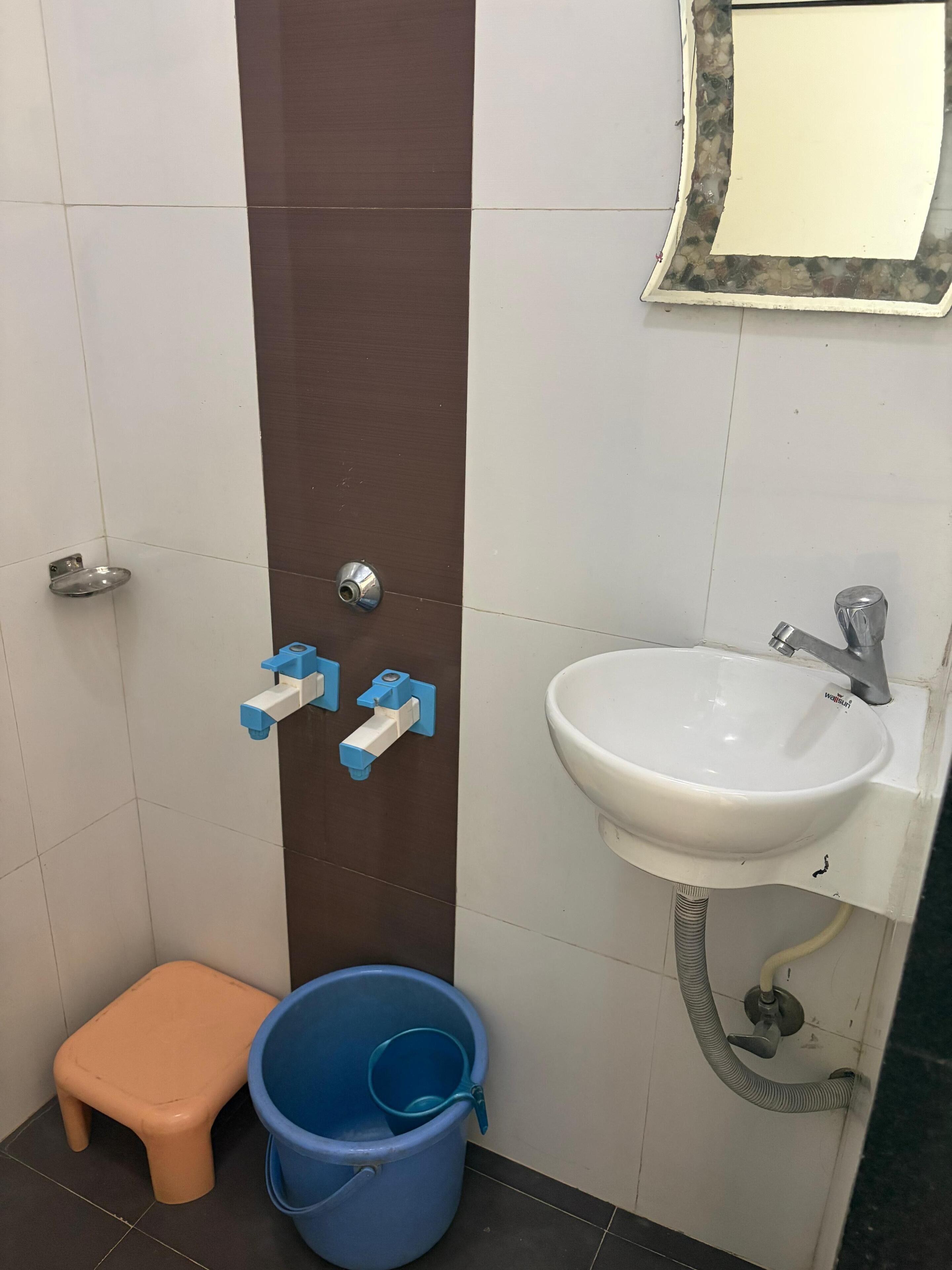 A bathroom with a white sink, a mirror, a brown door, a blue bucket, and a small orange stool.