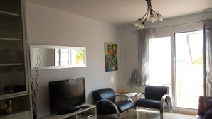 Ein modernes Wohnzimmer mit Flachbildfernseher, einer Couch, zwei Sesseln, einem Couchtisch und einem Essbereich mit kleinem Tisch und Stühlen.