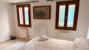 Ein Schlafzimmer mit Bett, zwei Fenstern, einem Heizkörper, einem Schrank und einem Bild an der Wand.