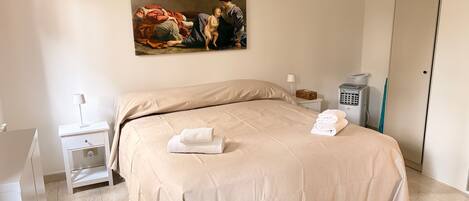 A hotel room with a large bed, a nightstand, a lamp, a trash bin, and a painting on the wall.