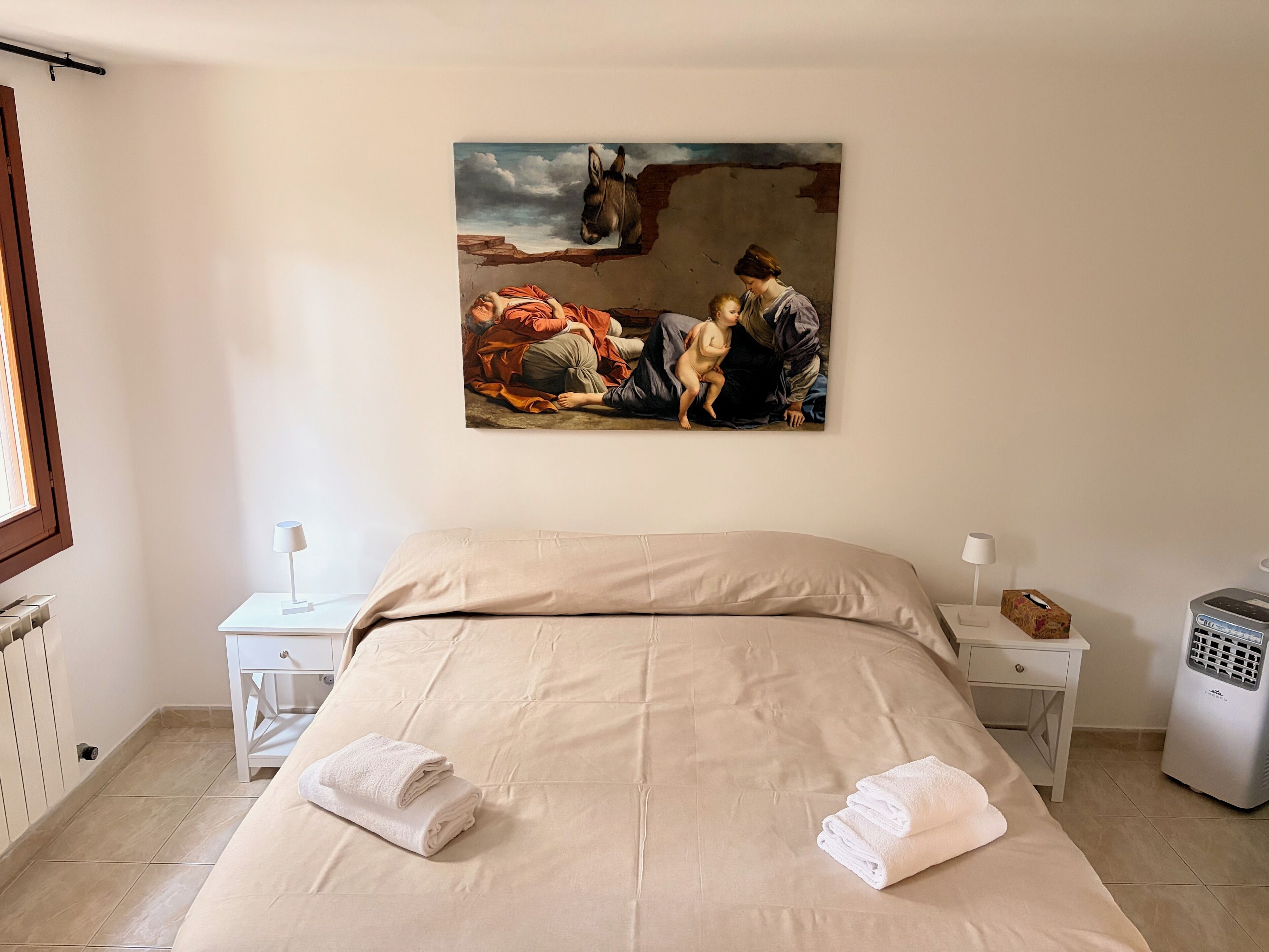 A hotel room with a large bed, two bedside tables, a radiator, a heater, and a painting on the wall.