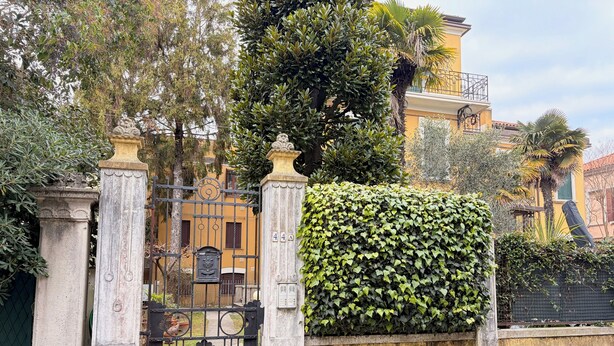 Ein verschlossenes Tor mit schmiedeeisernem Gitter, einer Steinpfeiler und einer Hecke.