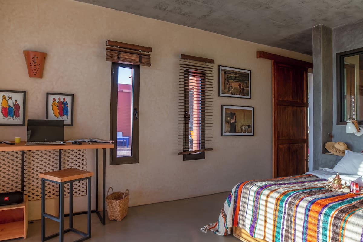 Una habitación con una cama, un escritorio con una computadora portátil, una ventana con persianas y una puerta.