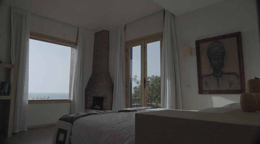 A bedroom with a fireplace, large windows, and a painting on the wall.