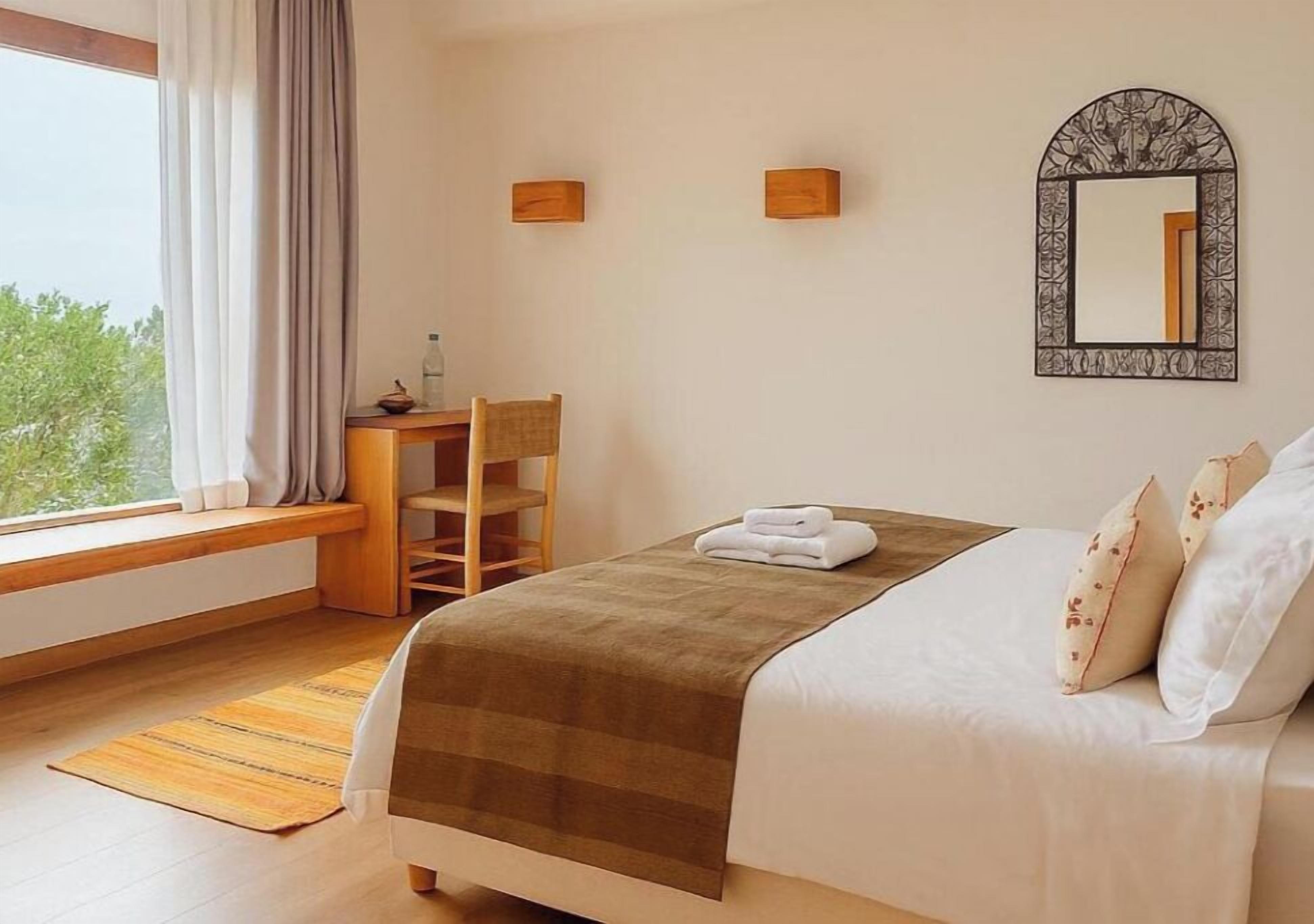 A bedroom with a bed, a desk, a chair, a window with curtains, and a mirror on the wall.