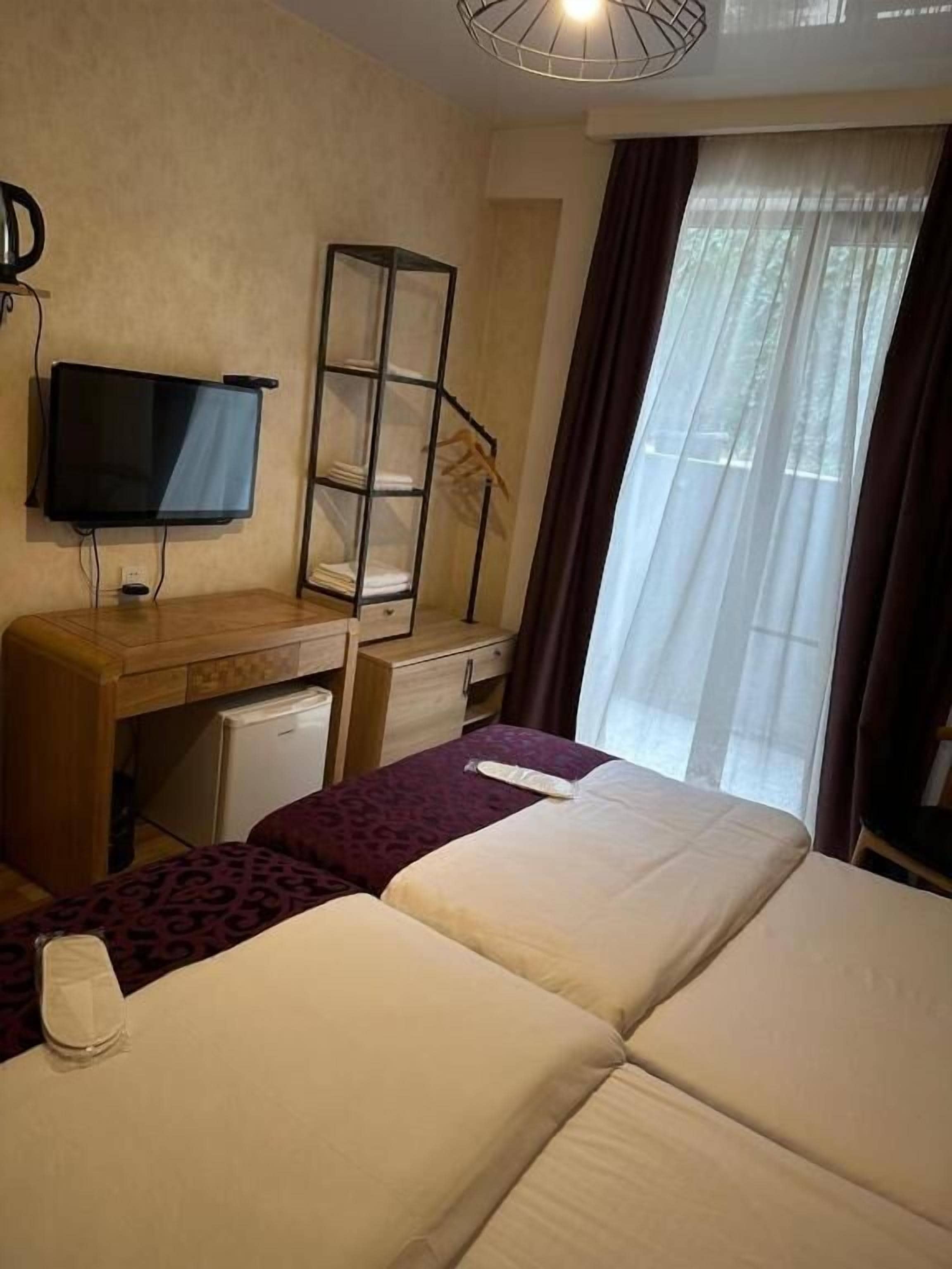A hotel room with a bed, a desk with a television, a wardrobe, and a window with curtains.