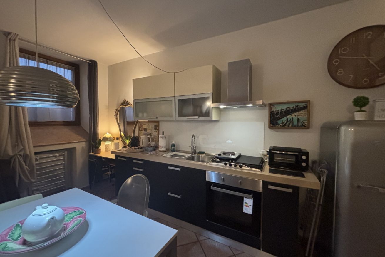 Una cocina compacta con estufa, microondas y lavamanos. Hay una mesa de comedor con una tetera y un plato decorativo. Un reloj está en la pared, y una planta sobre la encimera.