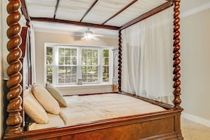 A four-poster bed with a wooden frame and a canopy, a window with blinds, and a ceiling fan.