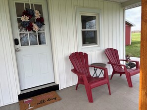 Una porta bianca con una ghirlanda, due poltrone Adirondack rosse, un tavolino e un tappetino d'ingresso con un bicchiere di vino.