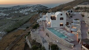 Ένα λευκό κτίριο με πισίνα και υπαίθριο χώρο για καθιστικό, περιτριγυρισμένο από πέτρινα τείχη και με θέα ένα χωριό στην πλαγιά του λόφου.