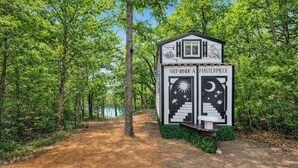 A cabin with a sign reading "Step Inside a Masterpiece" surrounded by trees and a dirt path. - The Doodle House | Lake Front Tiny Home (Bastrop County)