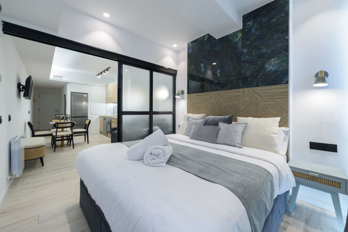 A modern hotel room with a large bed, a dining area, and a kitchen in the background.