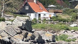Une maison avec un toit rouge, des murs blancs et des fondations en pierre, située près d’une étendue d’eau.