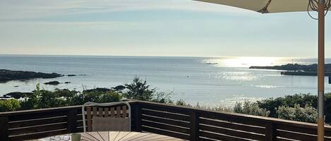 A wooden deck with a round table and chairs, an umbrella, and a view of the sea.