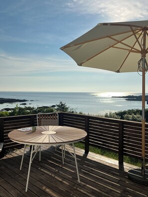 Een houten terras met een ronde tafel, stoelen, een parasol en uitzicht op zee.