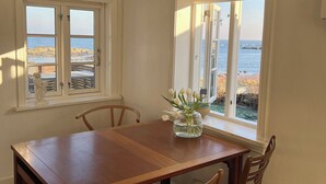 Une salle à manger avec une table et des chaises en bois, deux fenêtres donnant sur l’océan et un vase rempli de fleurs posé sur le rebord de la fenêtre.