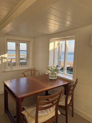 Comedor con mesa y sillas de madera, dos ventanas con vistas al océano y un jarrón con flores en el alféizar.