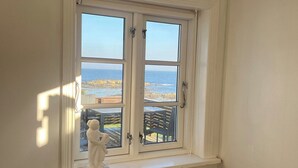 Une pièce avec une fenêtre blanche donnant sur une plage, une chaise en bois et une statue blanche posée sur le rebord de la fenêtre.